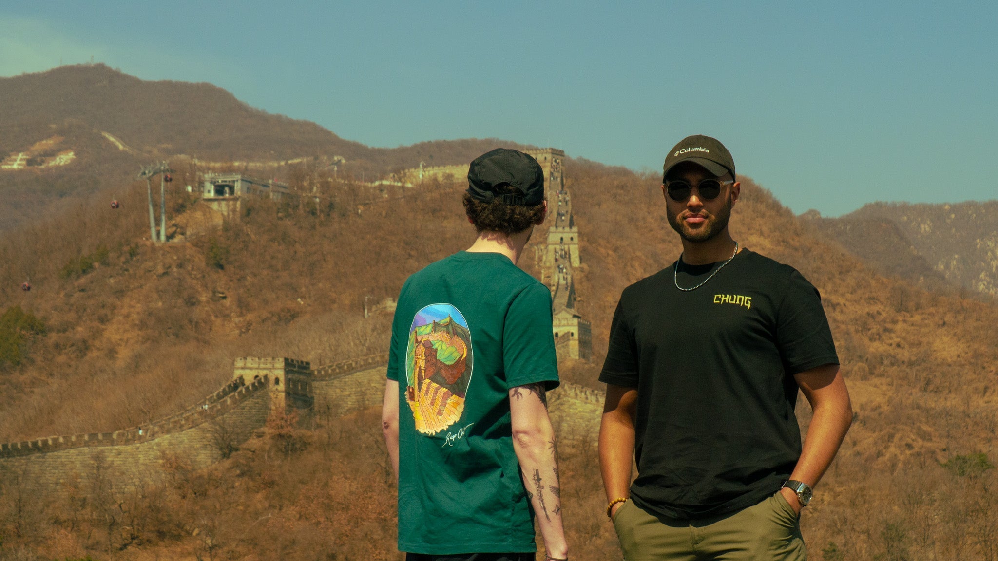 Two people standing on a mountain with a scenic view of a wall-like structure.
